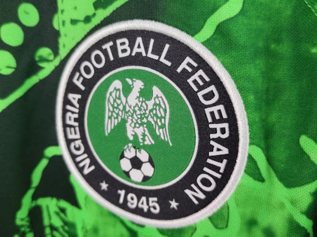 Gros plan sur l'écusson de la Fédération Nigériane de Football, représentant un aigle au-dessus d'un ballon de football, brodé sur un tissu vert à motifs graphiques noirs.