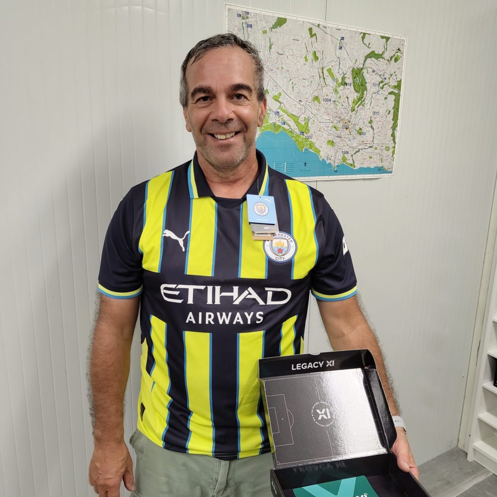 Un homme souriant porte un maillot de Manchester City et tient une boîte noire ouverte avec l'inscription "LEGACY XI"
